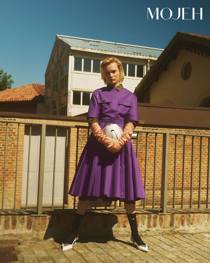 Model in a purple pleated dress and long gloves holding a white helmet outdoors against a brick railing.