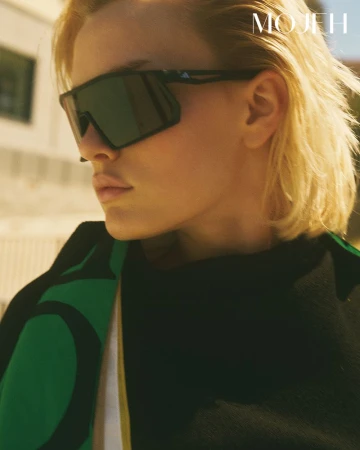 Close-up of a blonde woman wearing oversized black geometric shield sunglasses and a black garment with green details.