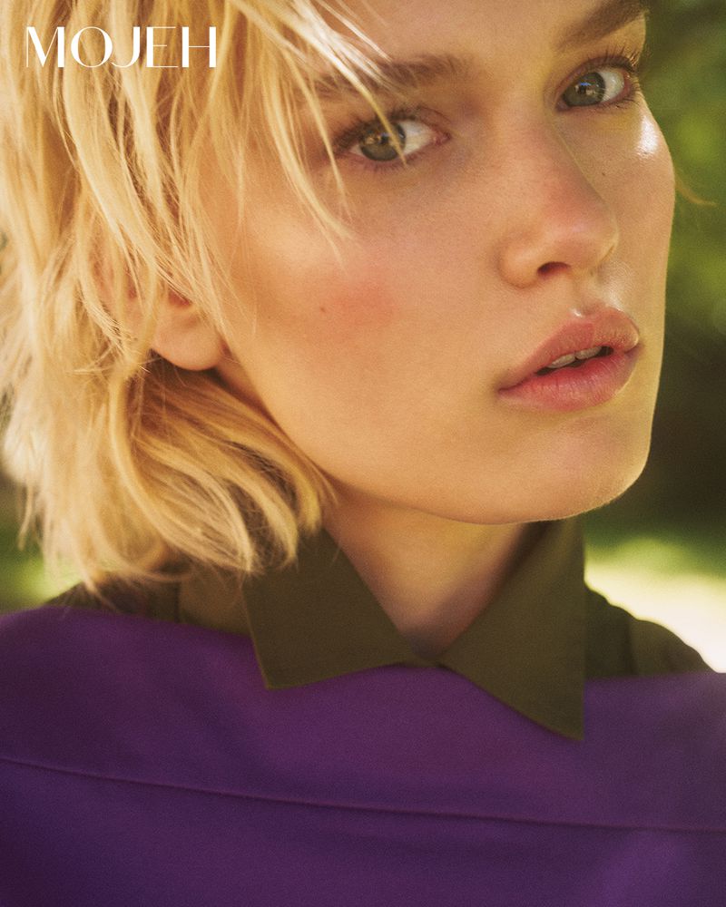Close-up portrait of a blonde female model with tousled hair, wearing a purple top and olive green collar, outdoors against foliage.