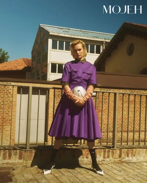Model in a purple pleated dress and long gloves holding a white helmet outdoors against a brick railing.