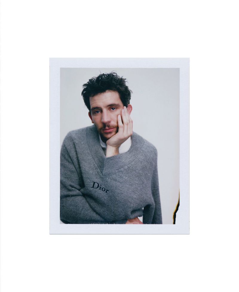 Josh O'Connor wearing a grey V-neck sweater with Dior branding, resting his chin on his hand in a portrait format.