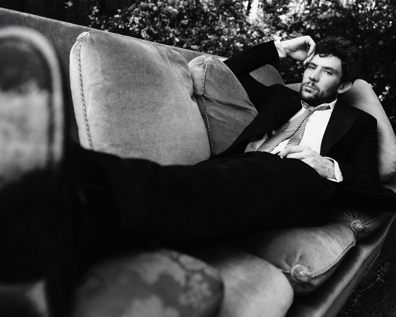 Actor Josh O'Connor reclining on a cushioned sofa outdoors wearing a suit, white shirt, and striped tie in black and white.