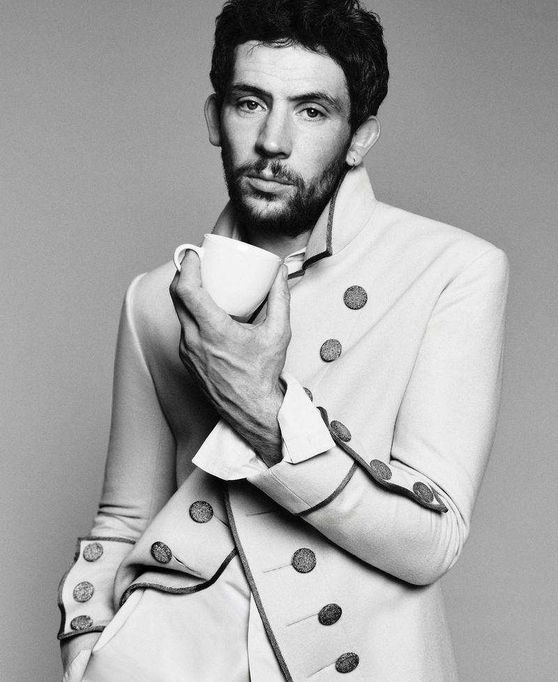 Actor Josh O'Connor holding a small white coffee cup while wearing a tailored military-style coat against a grey studio backdrop.