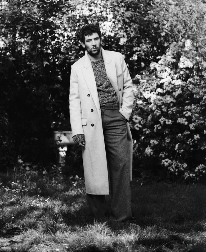 Black and white portrait of actor Josh O'Connor in a long tailored overcoat and knit sweater standing in a garden.