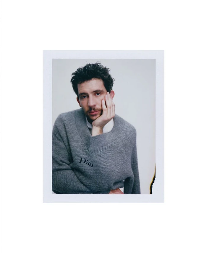Josh O'Connor wearing a grey V-neck sweater with Dior branding, resting his chin on his hand in a portrait format.