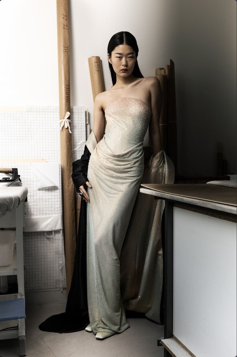 Model Yaxin Cao wearing a shimmering strapless champagne evening gown in a Paris fashion atelier workshop setting.
