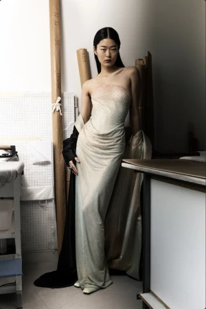 Model Yaxin Cao wearing a shimmering strapless champagne evening gown in a Paris fashion atelier workshop setting.
