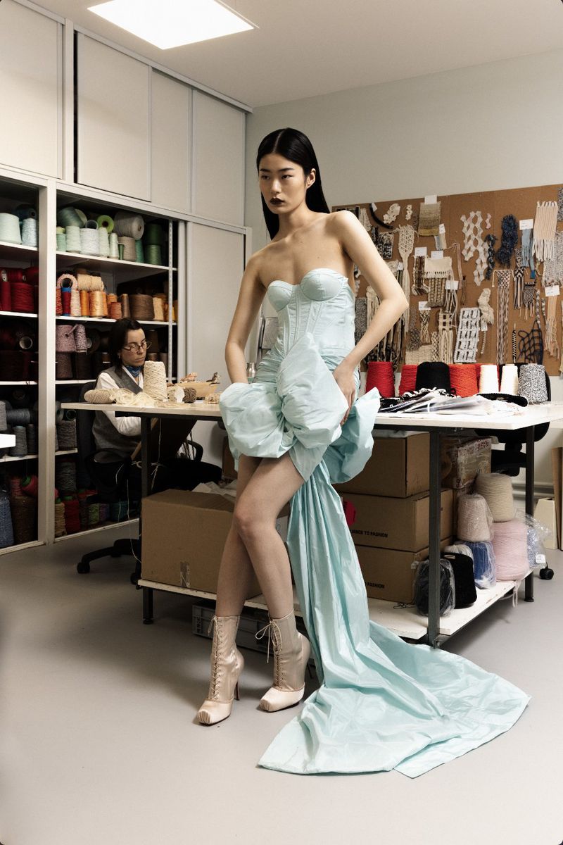 Model in a pale blue strapless dress with a dramatic train posing in a sewing workshop with shelves of yarn spools
