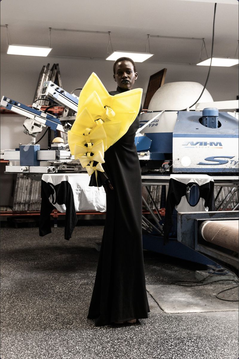 Model Toto Matoch in a floor-length black dress with an architectural yellow sleeve standing in a screen-printing atelier.