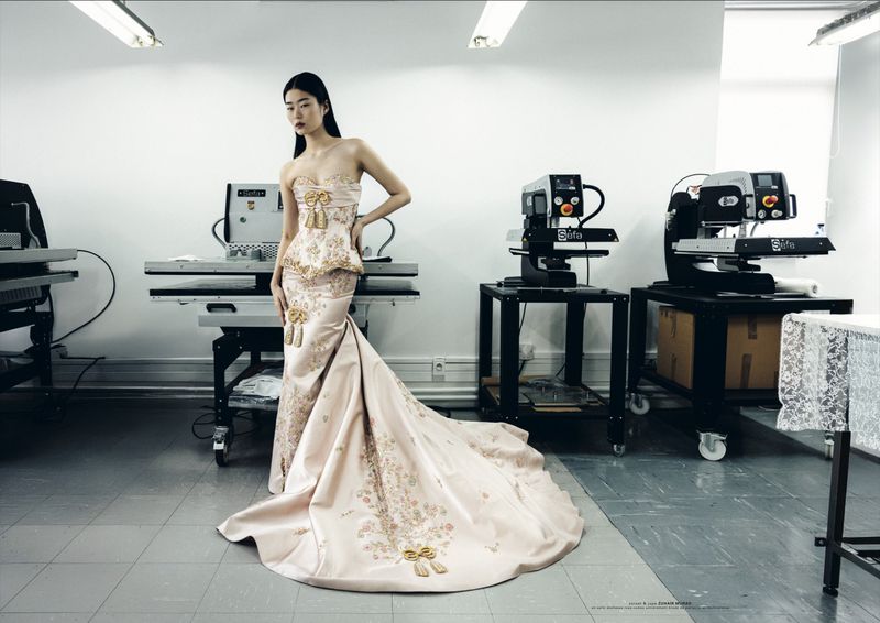 Model Yaxin Cao wearing an embroidered pale blush gown with a corset peplum and long train in an industrial atelier setting.