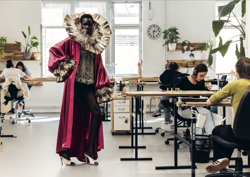 Model posing in an exaggerated gold ruff collar and deep magenta robe inside a bright Parisian fashion embroidery atelier.