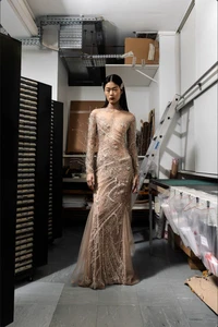 Model Yaxin Cao posing in a floor-length sheer embellished evening gown inside a workshop with storage racks and a ladder.