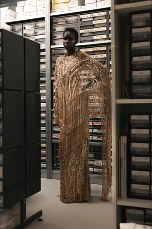 Model Toto Matoch wearing an ornate gold beaded fringe gown standing in a workshop storage room with component drawers.