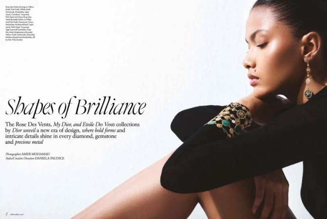 Brunette woman poses with gold Dior earring and bracelet with gemstones in a studio beauty shot, wearing a black long-sleeve shirt.
