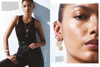 Model wearing layered gold necklaces, rings, earrings, and a black top against a white wall.
