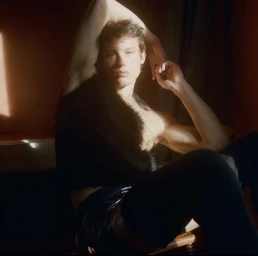 Model Eliott Tipp seated with his arm raised wearing a black textured vest in warm directional sunlight.