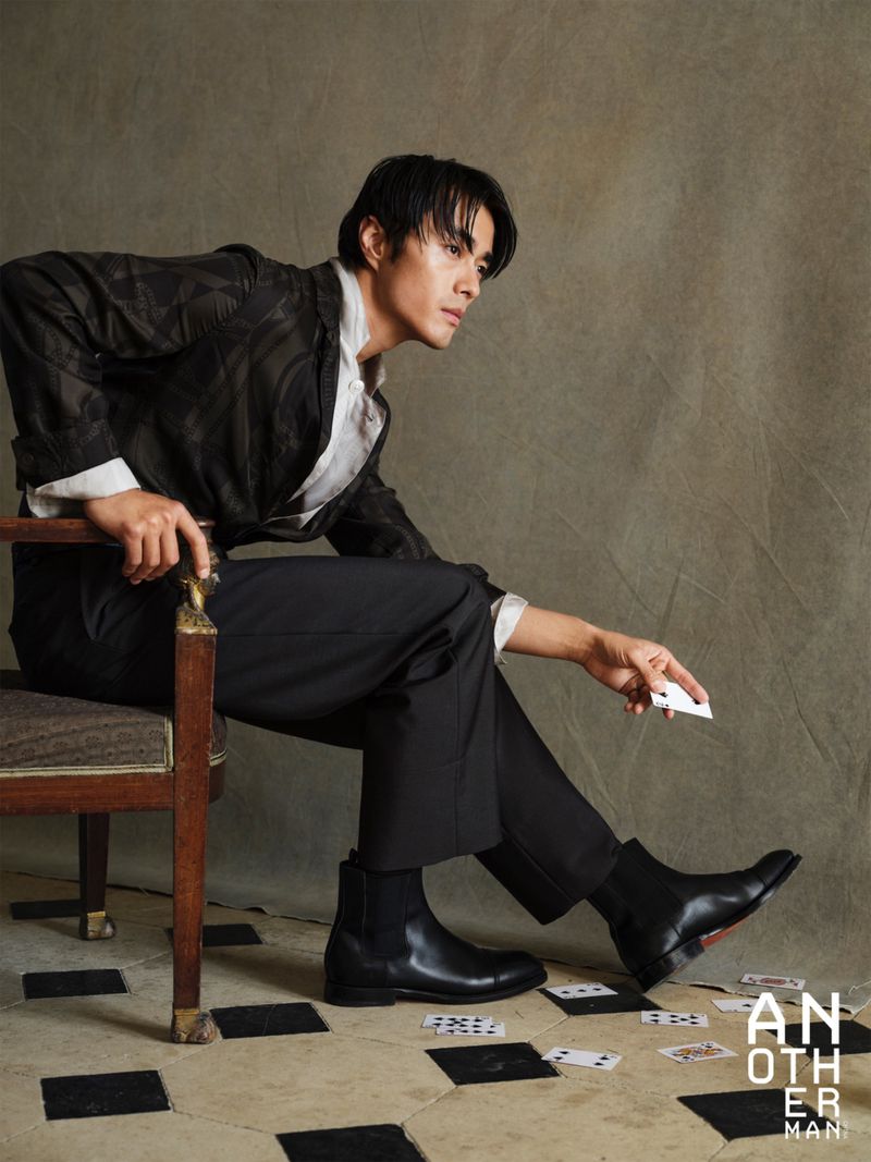 Model Zhao Lei wearing a dark jacket, black trousers, and leather boots seated on a vintage chair dropping playing cards.