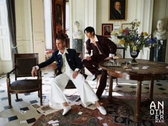 Two male models wearing tailored suits posed around a wooden table with a chessboard and scattered playing cards in a grand room.