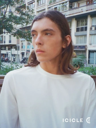 Model Ana Marín wearing a white long-sleeve crewneck shirt with an urban residential building in the background for ICICLE SS26 B Line.
