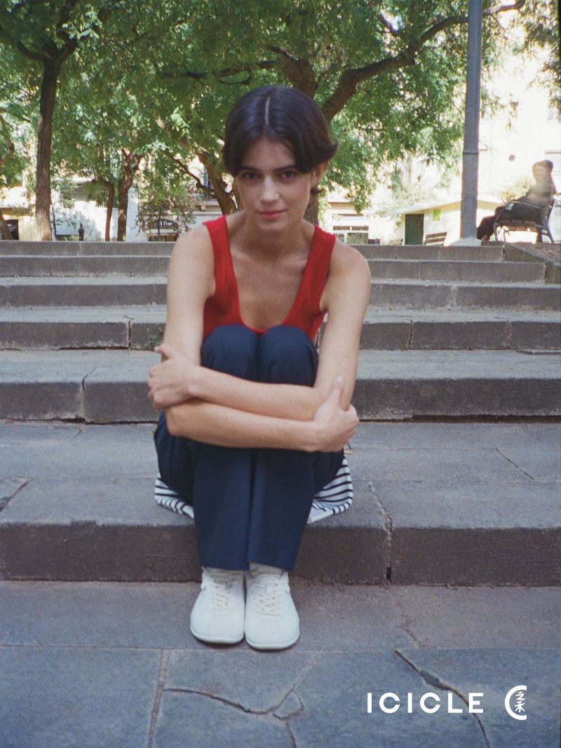 Model Ana Marín sitting on outdoor stone steps wearing a red tank top, navy blue trousers, and white sneakers.