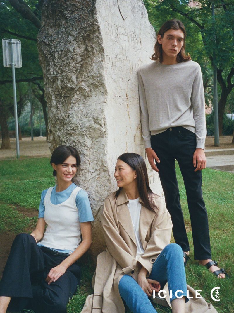 Three models wearing relaxed casual fashion posing by a large stone monument in a sunlit grassy park setting.