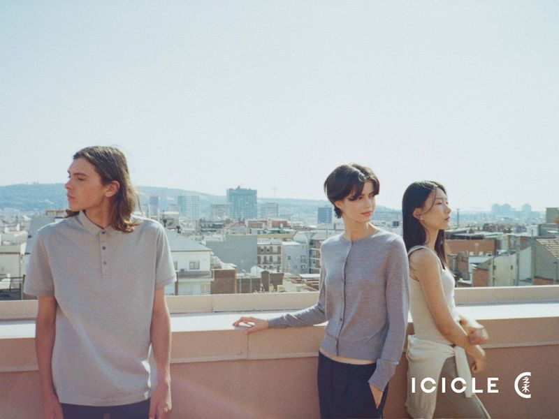 Three fashion models in neutral knitwear and polo shirts posing on an urban rooftop overlooking a cityscape.