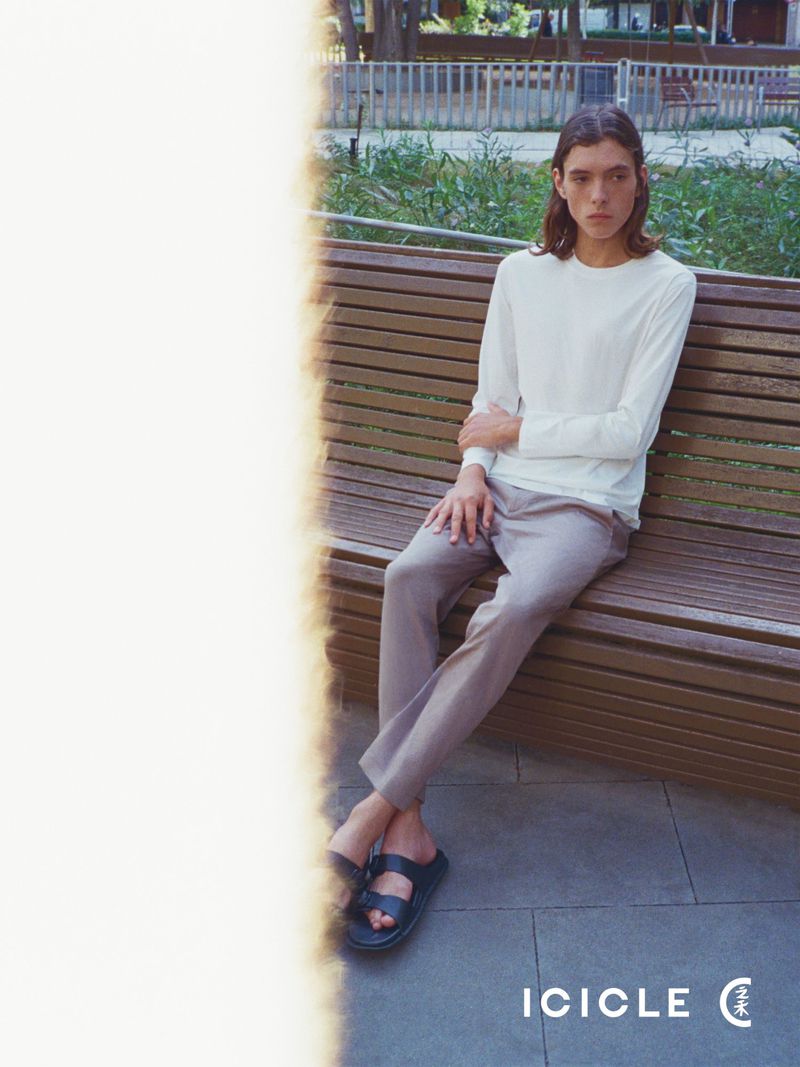 Model sitting on a wooden bench wearing a white long-sleeve top, taupe trousers, and black slide sandals in a park setting.