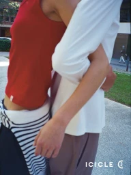 Close-up of two people embracing outdoors, one wearing a red tank top with a striped sweater tied at the waist, the other in a white top.