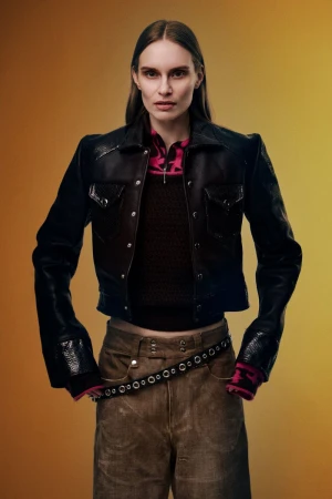 Model wearing a black cropped leather jacket and wide-leg brown trousers against a warm yellow-orange backdrop for REVS Magazine.
