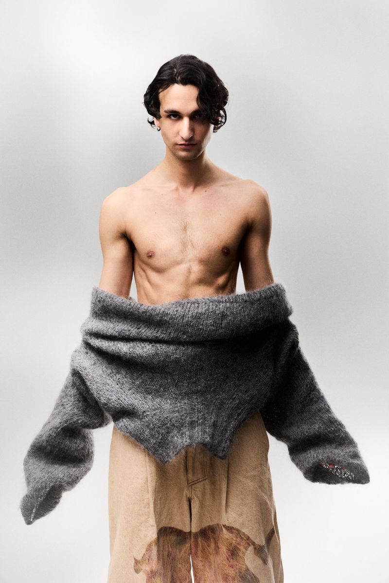 Shirtless male model wearing a grey knit sweater pulled down to his waist with printed beige trousers against a grey backdrop.