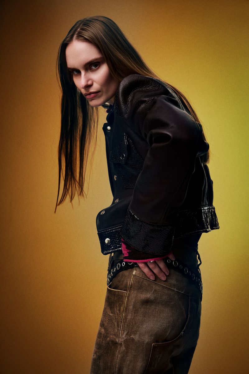Model in dark leather jacket and textured trousers posing against warm gradient background for REVS Magazine