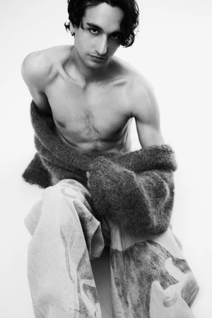 Shirtless male model posing in wide printed trousers and a dark fuzzy knit cardigan on a white background.