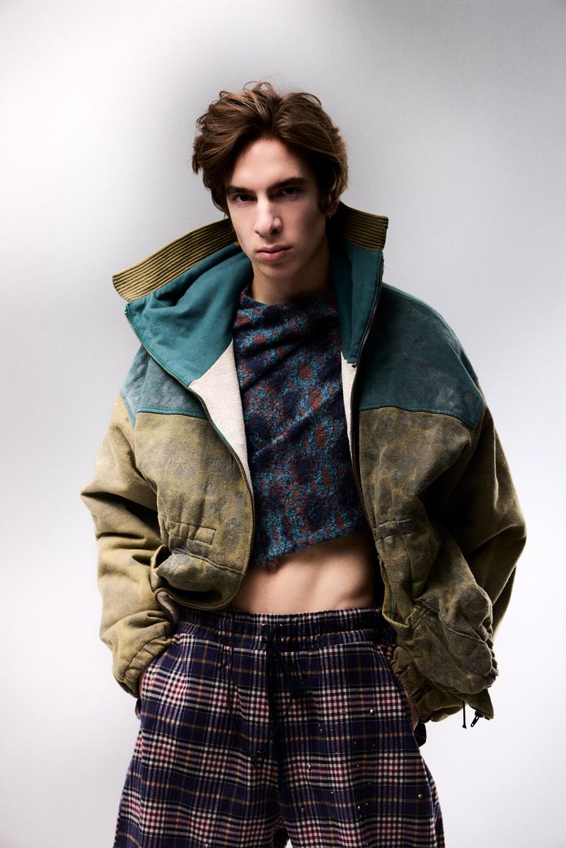 A young man in an oversized green jacket, cropped patterned top, and plaid trousers posing against a grey studio backdrop.
