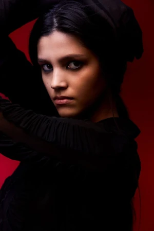 Portrait of a young woman with dark smoky eye makeup wearing a black ruffled garment against a deep red background.