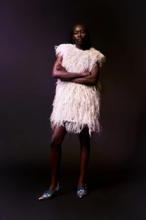 Fashion model wearing a textured white feathered mini dress with pointed silver heels against a dark seamless studio backdrop.