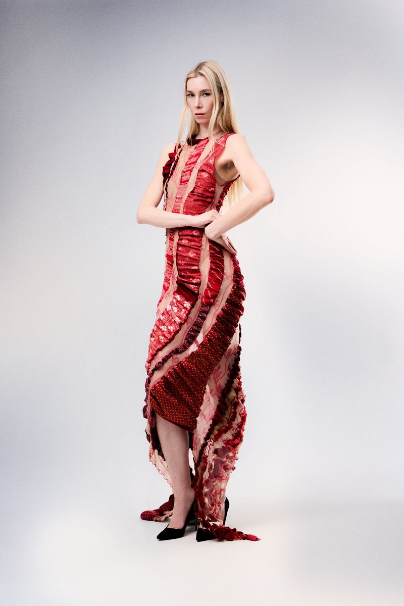 Blonde model in an intricate red ruffled sleeveless dress and black pointed heels posing against a grey seamless studio backdrop.