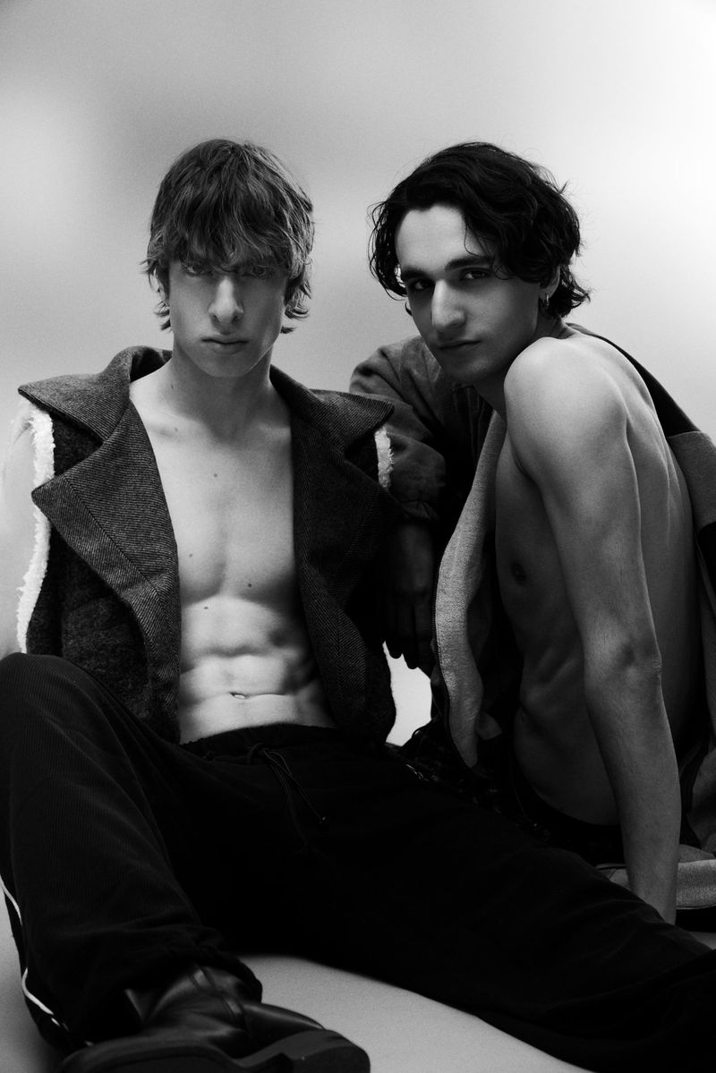 Two shirtless young men in dark shearling outerwear and trousers posing together against a studio background in black and white.