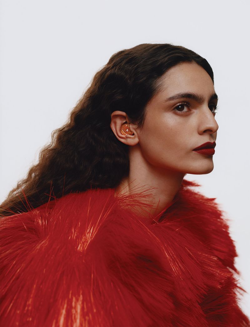 Model Carol Monteiro in a textured red feather coat and amber ear accessory against a light backdrop.