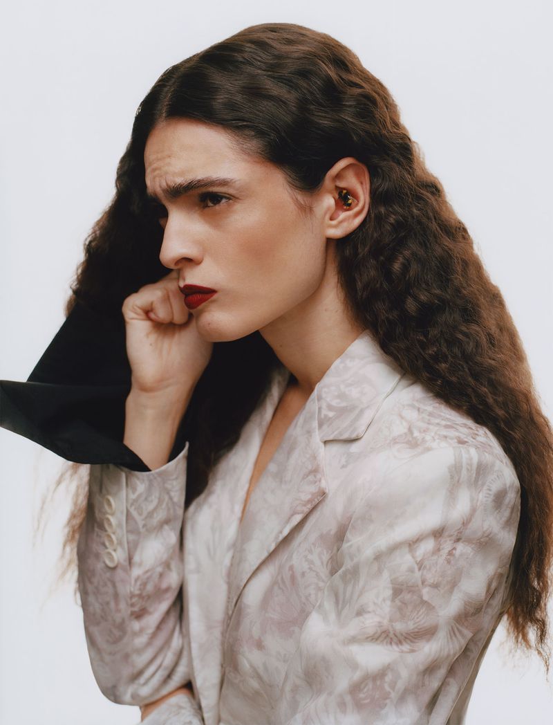 Model Carol Monteiro with long crimped hair wearing a pale jacquard jacket against a white studio background.