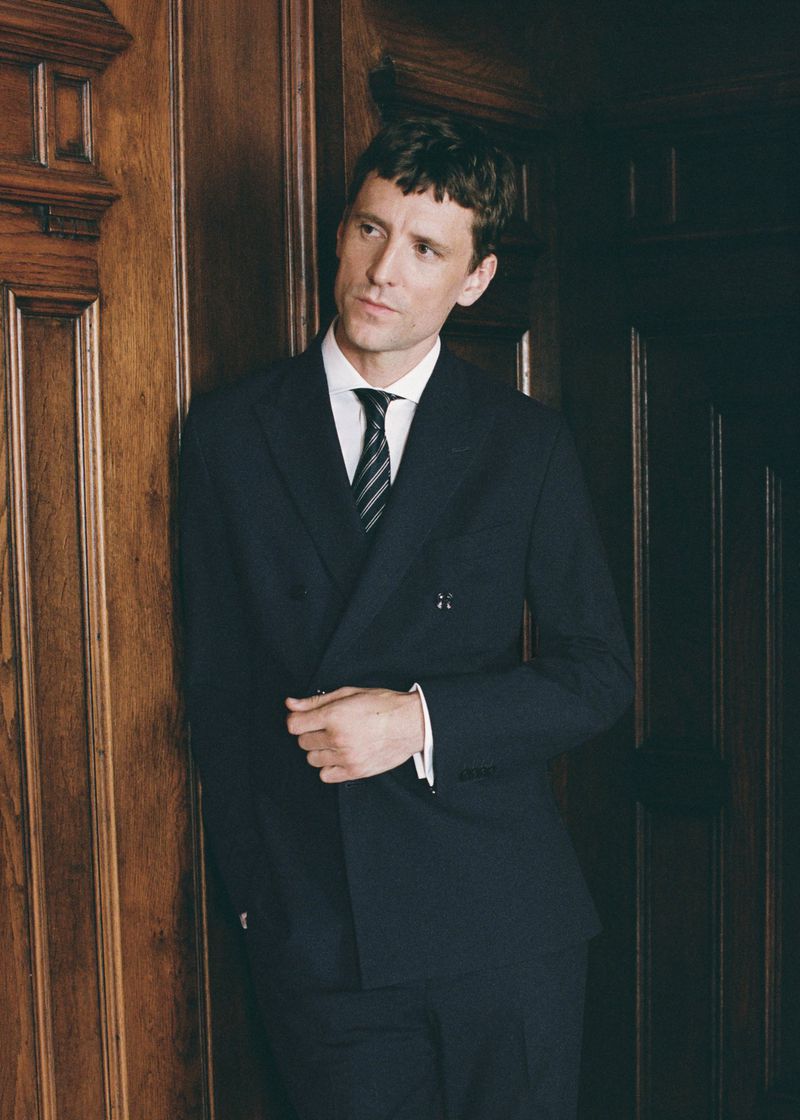 Model George Barnett leaning against wooden wall panelling wearing a dark double-breasted suit, white shirt, and striped tie.