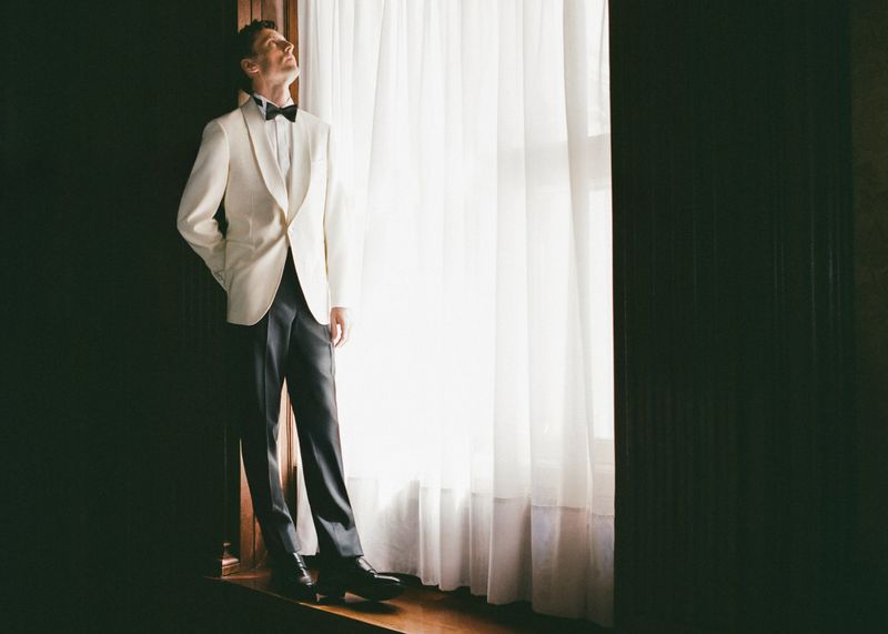 Model George Barnett in a cream tuxedo jacket, black bow tie, and black trousers stands by a window with white sheer curtains.