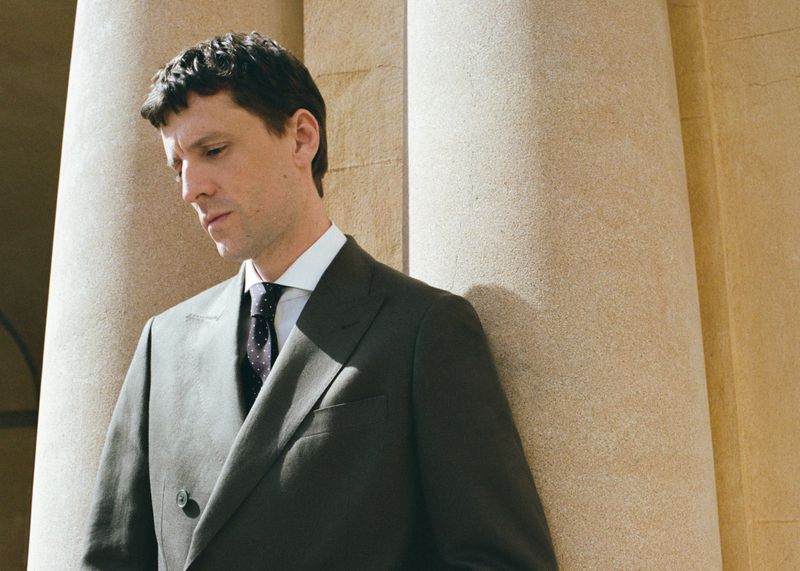 Man wearing a charcoal suit jacket, white shirt, and dark tie standing beside large stone architectural columns