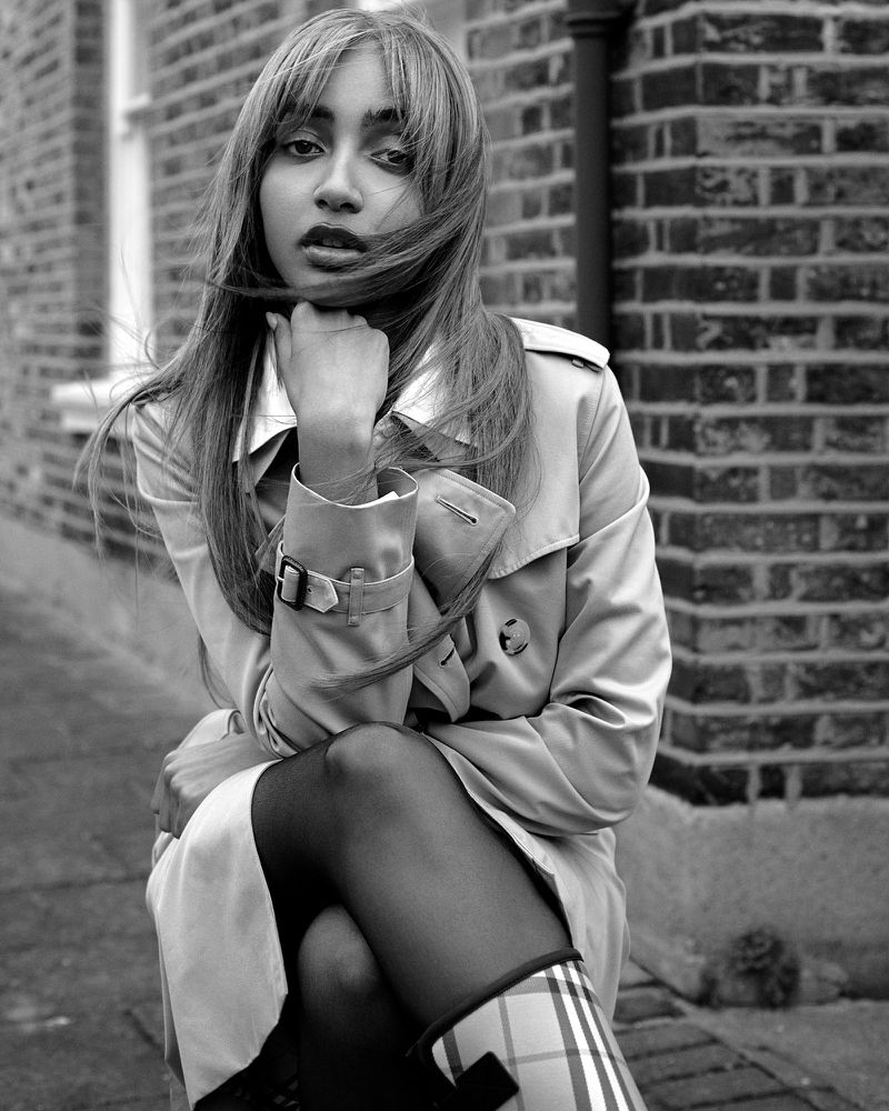 PinkPantheress sitting outdoors in a trench coat and check rain boots with her chin resting on her hand against a brick wall.