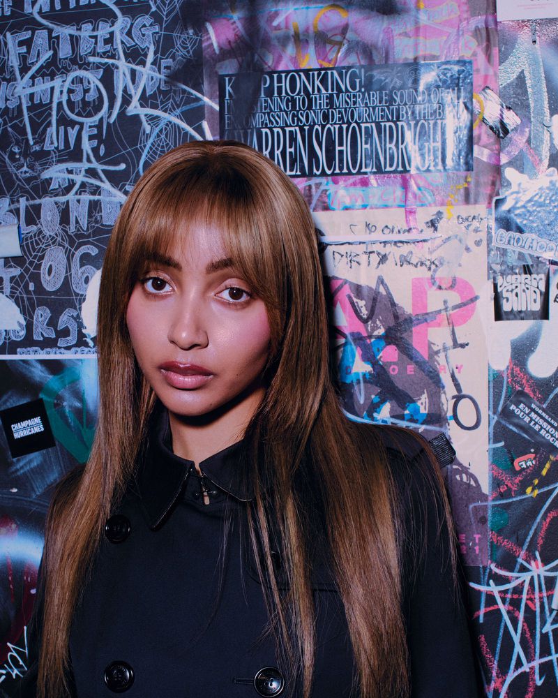PinkPantheress in a black collared coat standing before a wall covered in graffiti and posters.