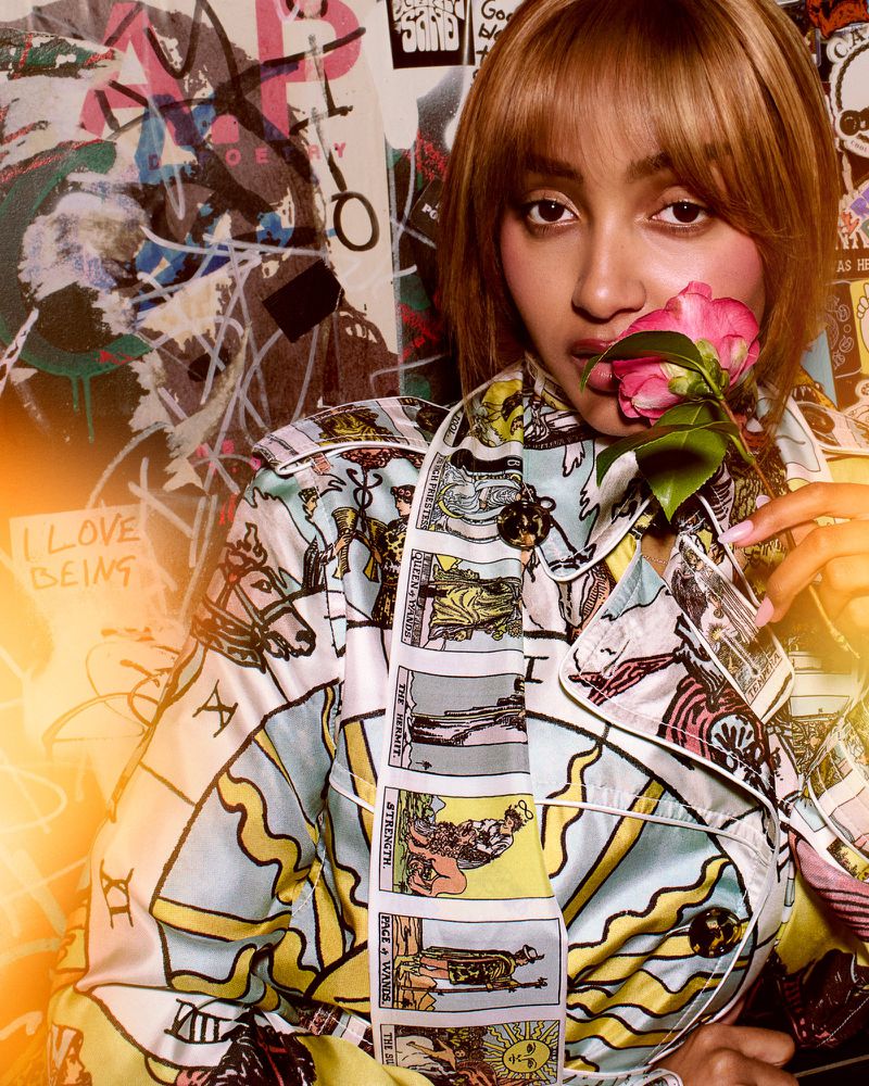 Close-up portrait of a woman holding a pink flower near her lips wearing a tarot card patterned coat against a graffiti wall