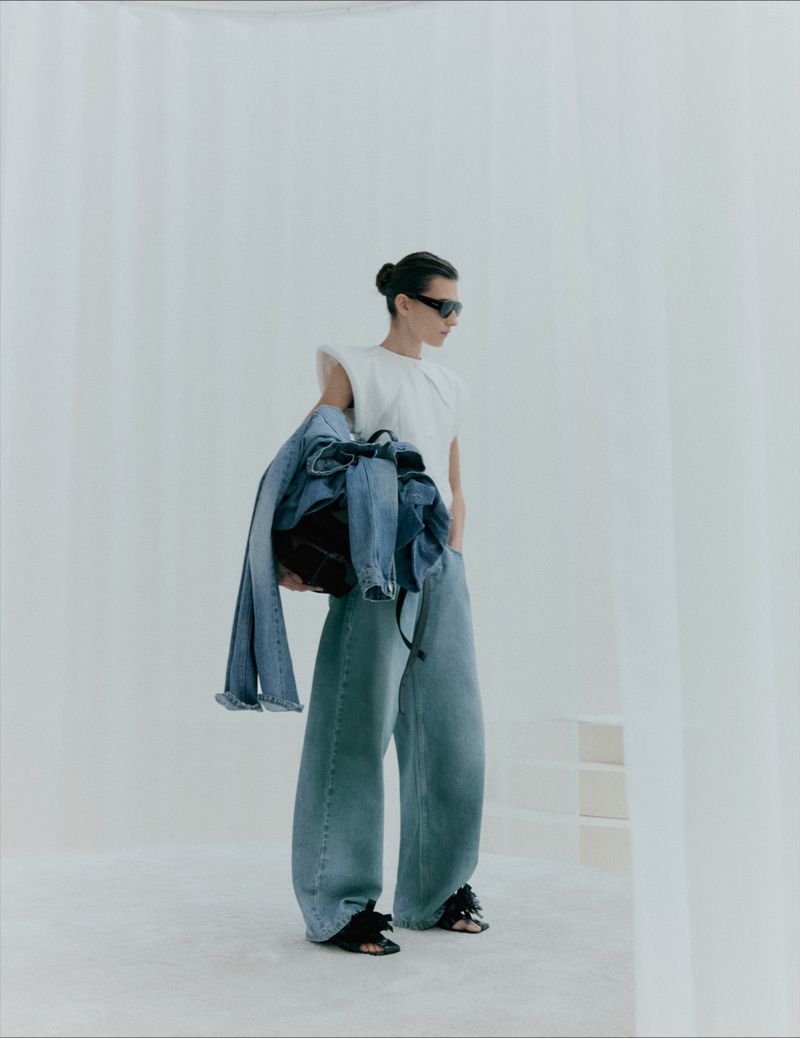 Model wearing wide leg denim jeans and sunglasses carrying denim jackets against a seamless white studio background