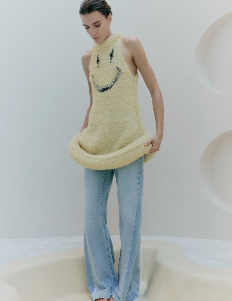 Model Irina Djuranovic wearing a fuzzy yellow smiley-face top and blue jeans against a minimal white background