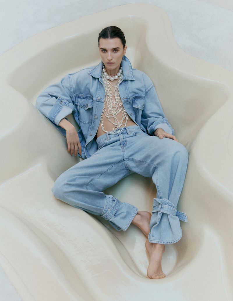 Irina Djuranovic wearing a light-wash denim suit and layered pearl necklaces, reclining in a sculptural beige seat for Vogue Scandinavia.