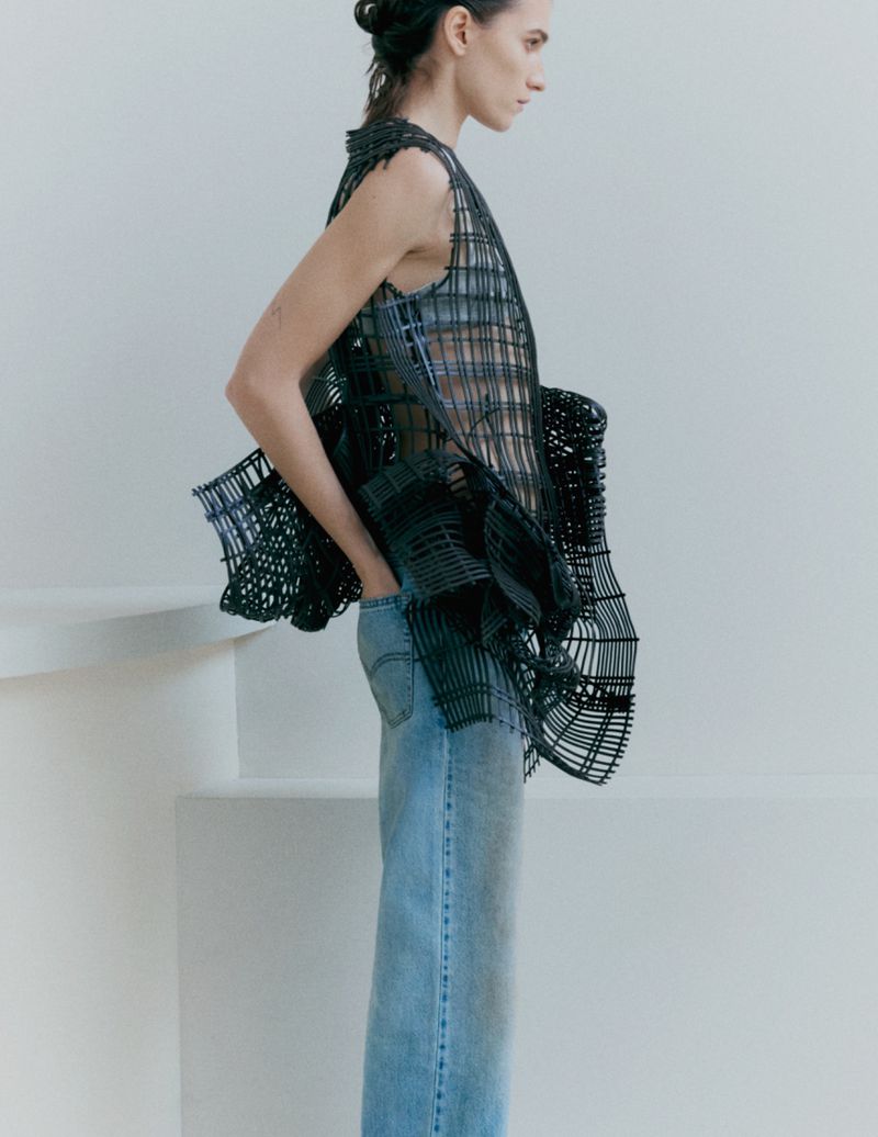 Model Irina Djuranovic wearing a black sculptural mesh top and light blue denim jeans against a pale white architectural studio backdrop.