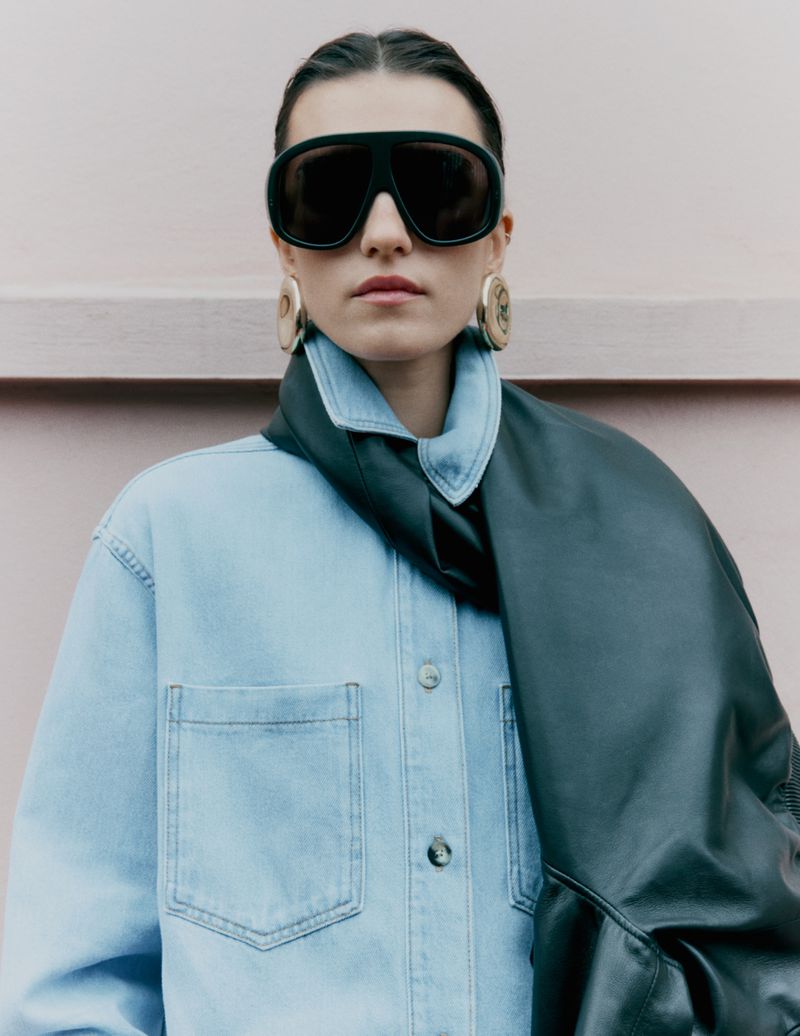 Model Irina Djuranovic wearing oversized black shield sunglasses and a light blue denim shirt against a pale wall.
